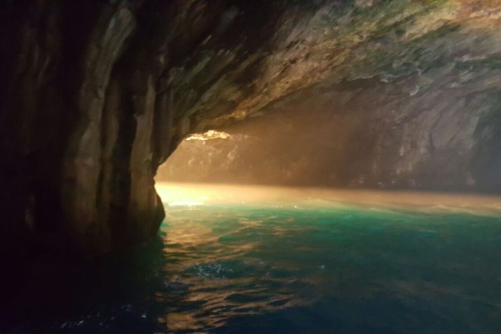 Sunlight illuminates the mouth of a rocky sea cave with blue-green water.