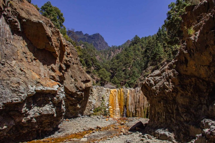 Rocky canyon with waterfall and forested mountains under clear blue sky.