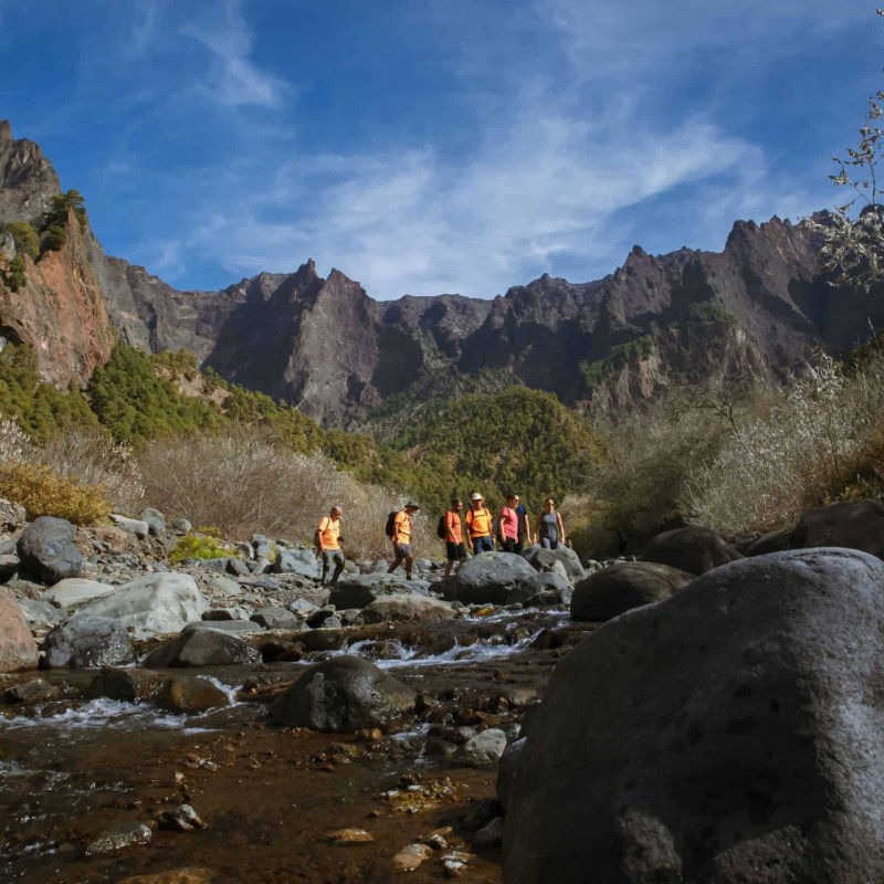 Group hiking in rocky valley with mountain backdrop under blue sky.