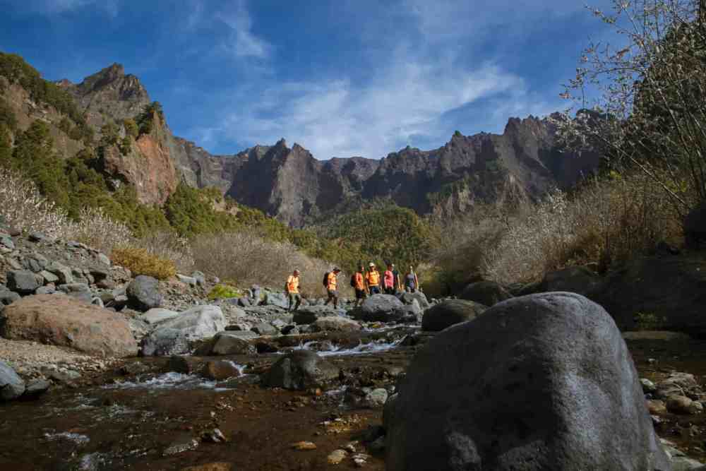 Hikers walking on rocky terrain beside a mountain stream under a blue sky.