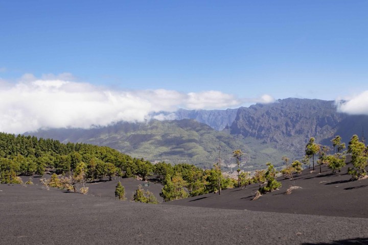 Mountain landscape with black sand, scattered trees, and clouds under a blue sky.