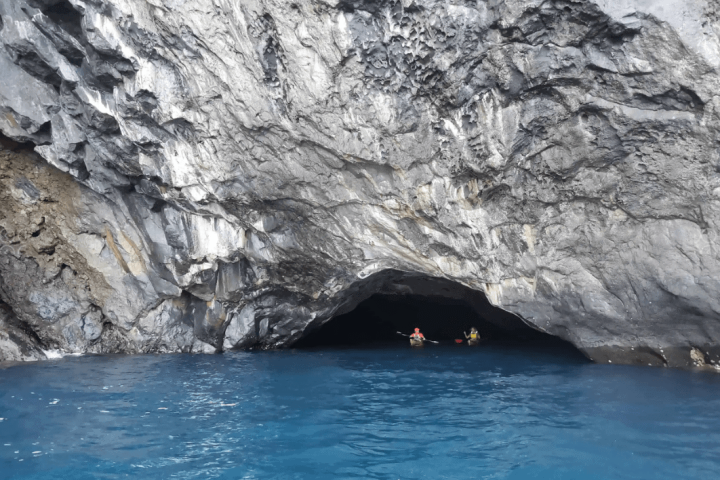 Kayakers near a rocky cave entrance above blue water.