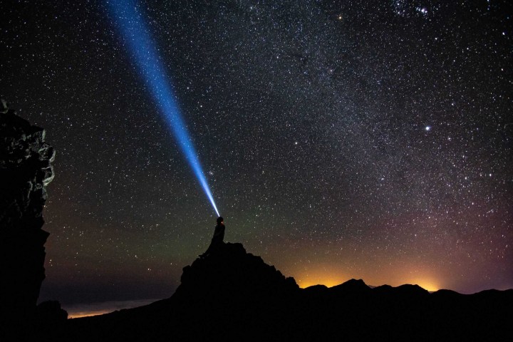 Silhouette on hill shines flashlight into starry night sky.