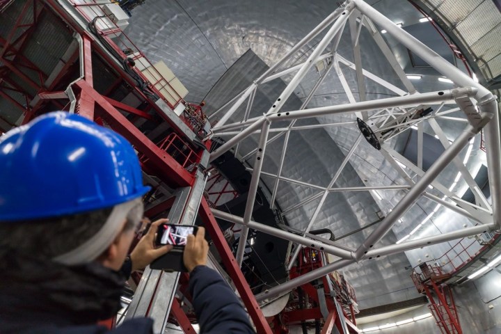 Person in blue helmet photographing large telescope structure indoors.